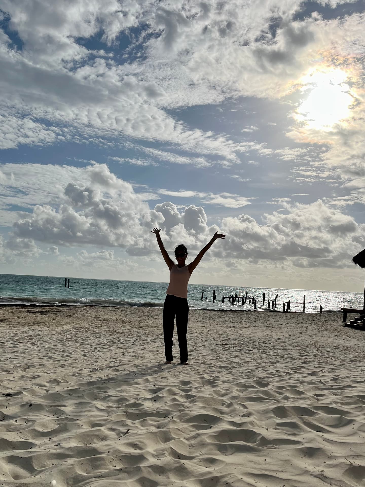 Standing on a beach with arms raised, backlit by the sun, old wooden pilings in the surf behind